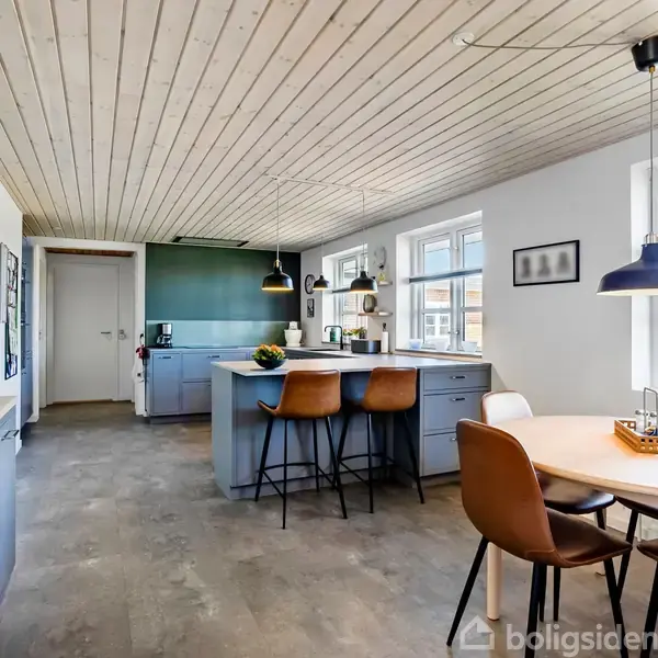 Et moderne køkken med blå skabe og en central ø, hvor to høje stole står. Rundt spisebord med fire stole. Rummet er lyst med store vinduer og lamper i loftet.