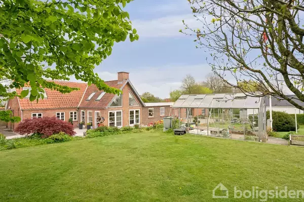 Et rødt murstenshus med tegltag står i en grøn have ved siden af et glashus. Haven har græsplæne og træer under en blå himmel.