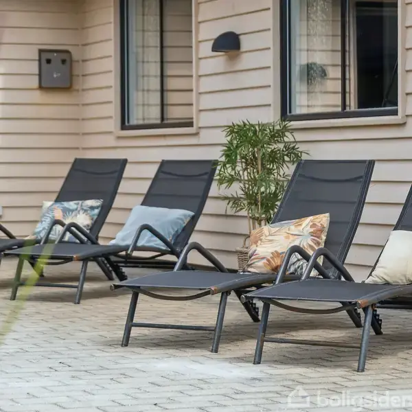 Et moderne sommerhus med fladt tag står på en åben græsplæne. Det har en terrasse med havemøbler og en grill. En grøn rutsjebane er ved siden af terrassen.