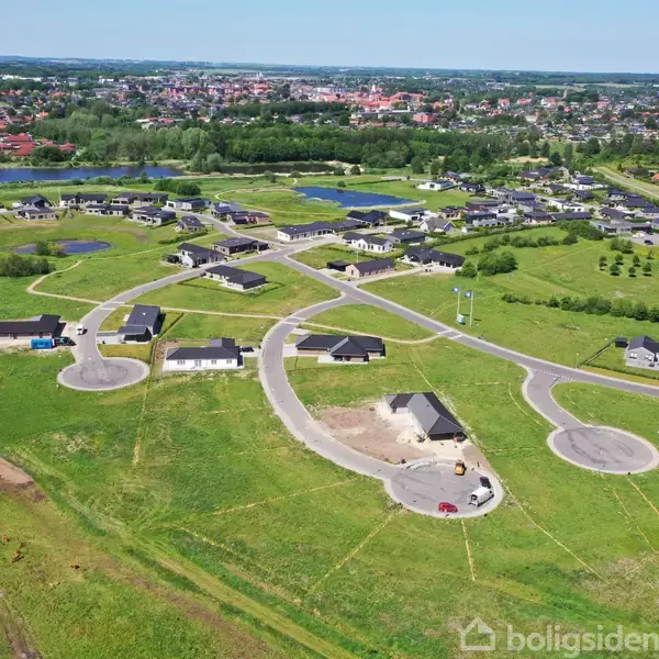 Nye huse under opførelse ligger spredt på grønne græsarealer, hvor veje forbinder dem. Området strækker sig mod en by i baggrunden med søer og flere huse nær horisonten.