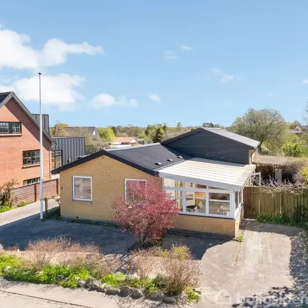 En gulstensvilla med sort tag står ud mod vejen. Foran er en indkørsel og have. Villaen ligger i en villavej omgivet af træer og andre huse i et grønt nabolag.