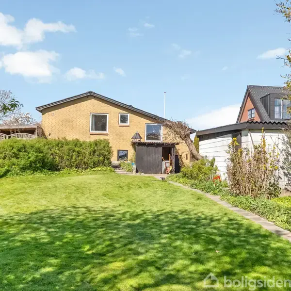 En gul murstenshus med træåbninger står i forgrunden. Huset har skrå tag, omkranset af frodig have med grøn græsplæne, buske og et træ. Solrigt vejr med blå himmel.