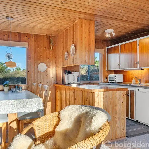 Køkken med træpaneler. Bord med dug står ved vindue. Skabene er træ, og der står køkkenudstyr. En rattanstol med fåreskind er i forgrunden, omgivet af naturligt lys.