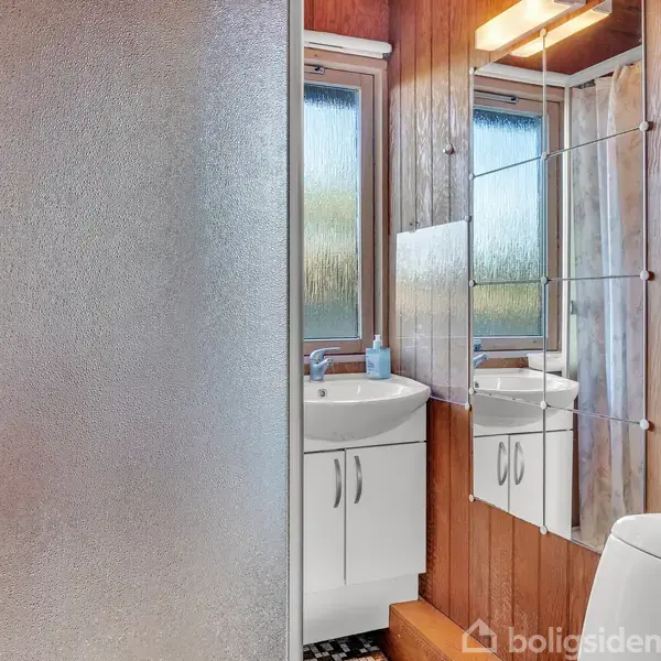 Et badeværelse med trægulve har et bruseafsnit med matteret skillevæg. Vaskemaskinen står under et vindue, og et spejl hænger over et skab med to skabe.