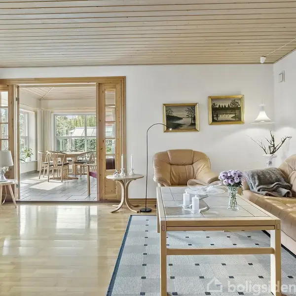 Brunt lædersofa-sæt står omkring et sofabord med blomster. Rummet har lyse trægulve, vægge med malerier, og store vinduer, der lader lyset strømme ind fra den tilstødende spisestue.