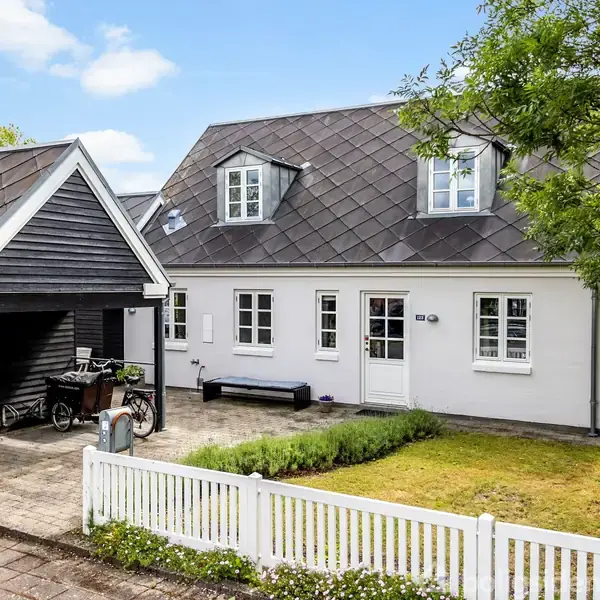 Hvidt hus med sorte vinduer og dør står ved siden af en garage med cykler. Foran huset er en lille græsplæne omgivet af et hvidt stakit.