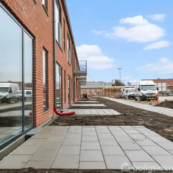 Rækkehus i rød mursten står langs en gangsti med fliser. Adskillige biler er parkeret nær bygningen. Det åbne område er under opbygning med jord og byggematerialer omkring.