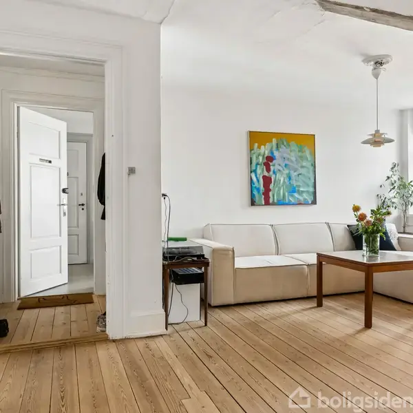En lys stue med en beige sofa ved væggen. Ovenfor hænger et farverigt maleri. På sofabordet står en vase med blomster. Et stort vindue giver udsigt til himlen.