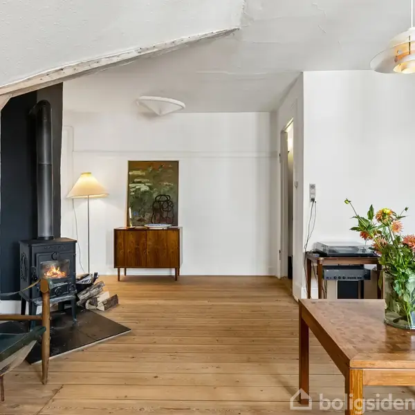 Stue med brændeovn i hjørnet, træbord i forgrunden med blomster, lænestol til venstre, og en lampe ved et skab i baggrunden. Hvidmalede vægge og trægulv.