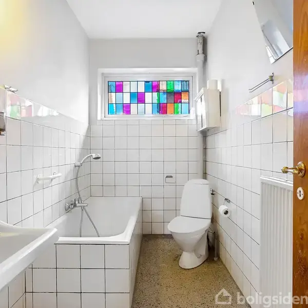 Badeværelse med hvidt flisebelagt badekar og håndvask, toilet ved siden af. Farvet glas i vinduet over toilettet. Lyse fliser dækker væggene; gulvet er gråt med pletter.