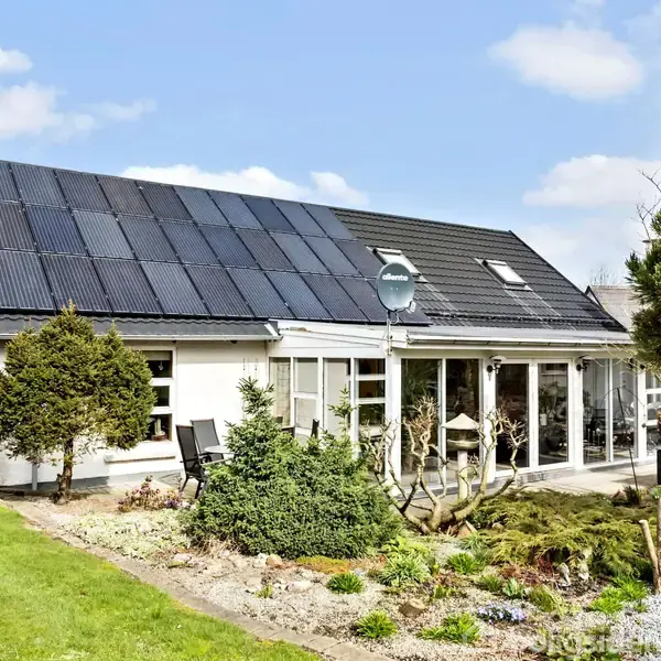 Hus med solpaneler på taget, omgivet af en have med buske og træer, under en blå himmel med få skyer. En satellitdisk er monteret på taget.