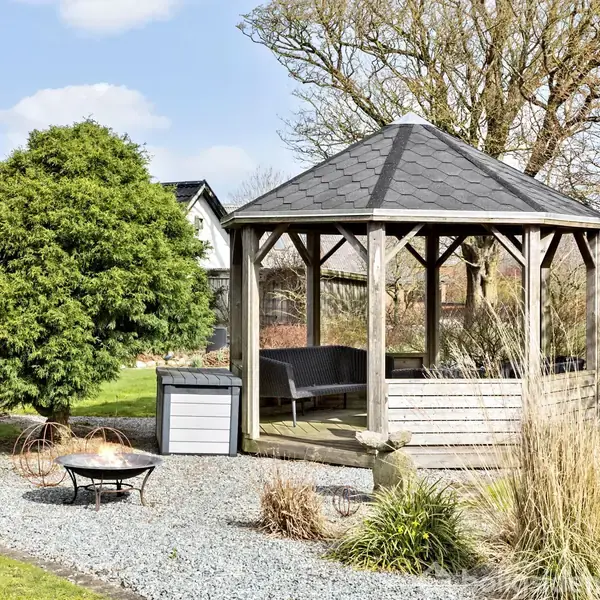 Pavillon står stille på en grusbelagt overflade omgivet af grønne træer og buske. Det er en åben trækonstruktion med tag, og i nærheden står et bålfad.