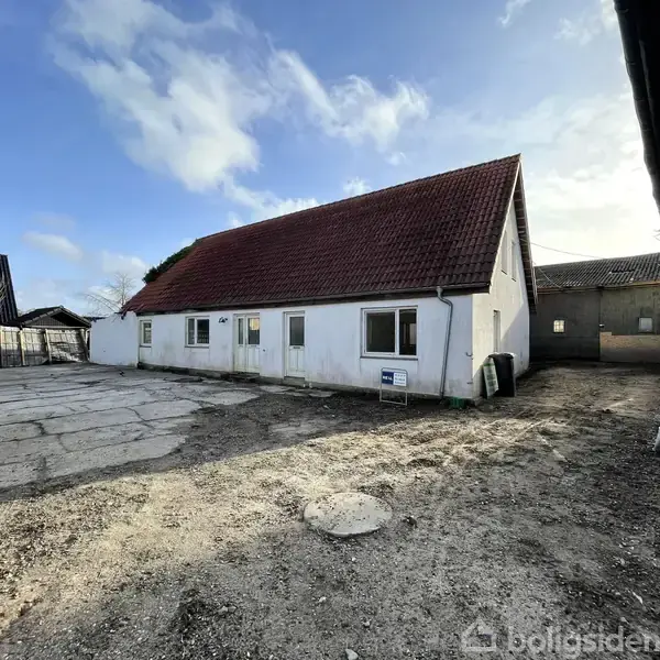 Hvidt, enkelt hus med rødt sadeltag står i en gårdsplads under en blå himmel. En skilt med teksten "Boligsiden" står ved siden af huset. En stor åben grusplads omgiver bygningen.