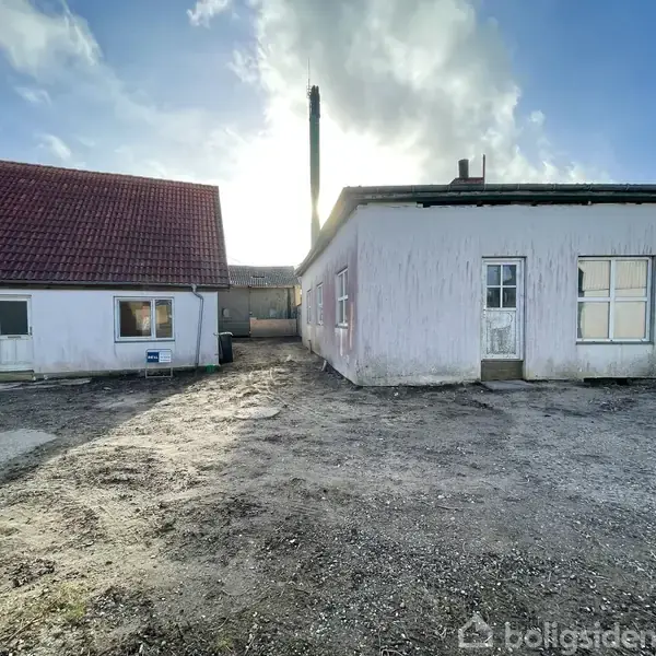 To bygninger stående på en grusbelagt gårdsplads, én med rødt tag og en enkelt skorsten stikkende op. Himlen er delvist dækket af skyer, og solen skinner bag bygningerne.