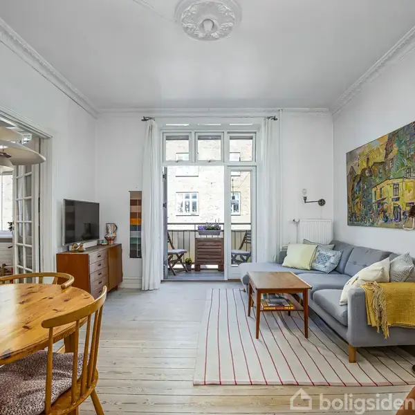 Stue med grå sofa, sofabord og tæppe, beliggende ved siden af altandør. Rundt bord til venstre med blomster. Hylder og fladskærms-tv til højre. Lyse vægge og kunst på væggen.