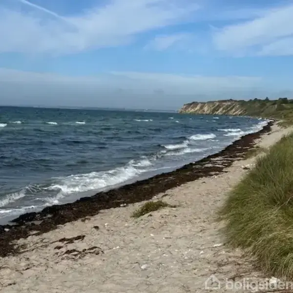 Kystlinje strækker sig langs en bølgende havoverflade med blåt vand. Sandstrand dækket af tang ligger mellem klitter og havet, mens klipper ses i horisonten under en blå himmel.