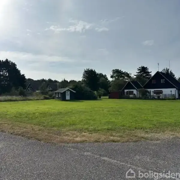 Grønt græsareal ligger stille foran sorte og hvide huse på en fredelig dag, omgivet af træer og blå himmel.