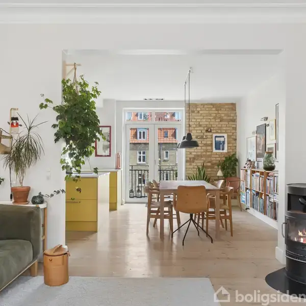 Sofa med grøn farve står i stue med planter og brændeovn. I baggrunden ses spisebord og stole ved vindue mod murstensvæg. Værelset er lyst og moderne.
