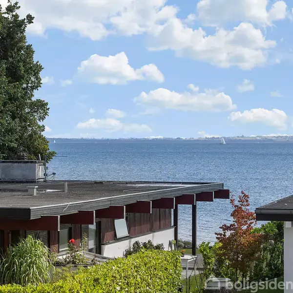 Et hus ligger ved kysten, med en flad tagkonstruktion. I baggrunden ses havet, hvor en sejlbåd glider gennem vandet under en blå himmel med spredte skyer.