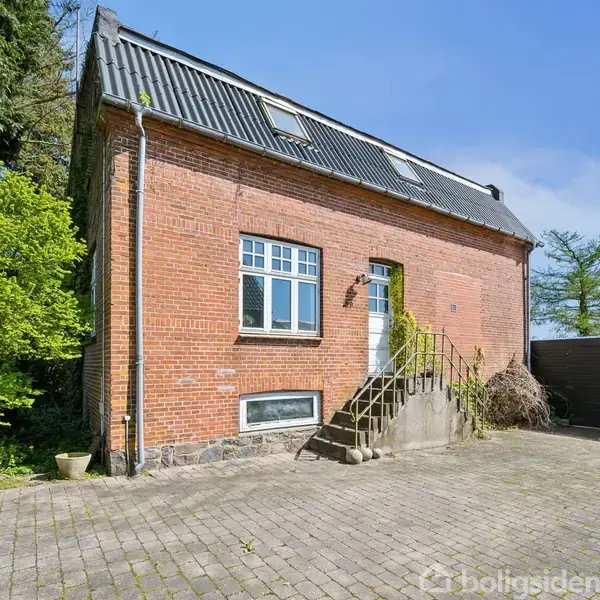 En rød murstensbygning med skråt sort tag, to vinduer og en hvid dør med trappe. Omgivet af flisebelagt gårdsplads og grønne træer.