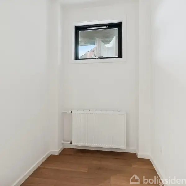 Et lille rum med hvide vægge, trægulv og en radiator under et lille vindue. Vinduet giver et begrænset udsyn til det udendørs område.