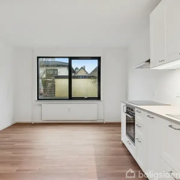 En hvid treetagers bygning med sorte døre og vinduer står stille i et gårdmiljø med asfalt og en hvid bil foran. Bygningen har altaner og kvistvinduer.