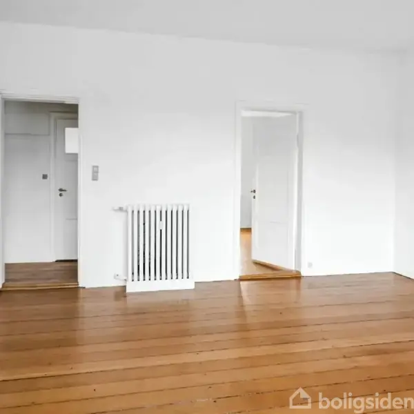 Et tomt rum med lyse træplanker på gulvet. To åbne døre fører til andre rum, og en radiator hænger på væggen. Væggene er hvide.