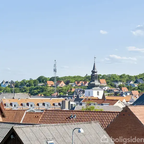 En kirke med et spir står blandt hustage. I baggrunden ses en grøn bakke dækket med træer og flere huse. Himlen er klar og blå.