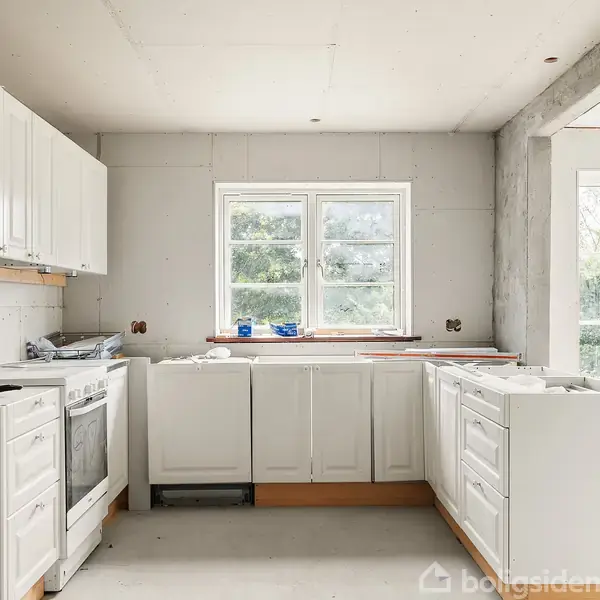 Lyst køkken under renovering med hvide skabe, en ovn og åbne hylder. Et stort vindue viser grønne træer udenfor. Golvet og væggene ser ufærdige ud.