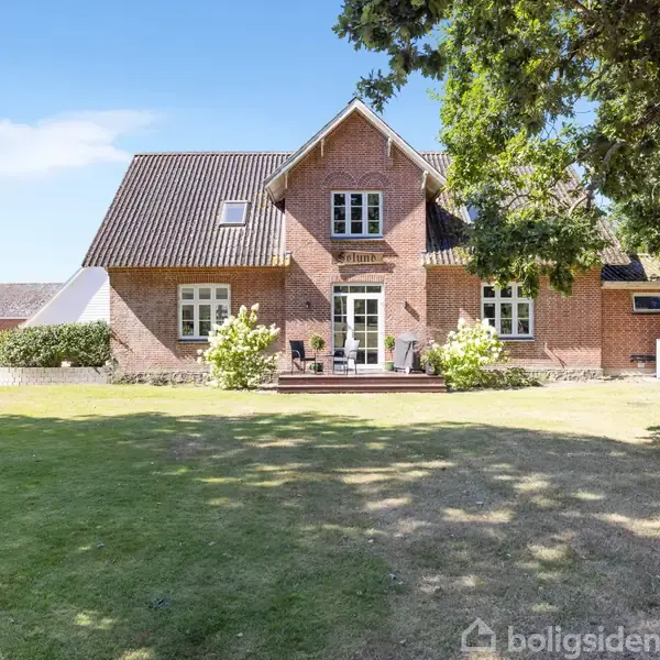 Hus med rødt murværk står i solrig have med grøn græsplæne. Et stort træ kaster skygger til højre. Skilt over døren siger "VILLADOLF."