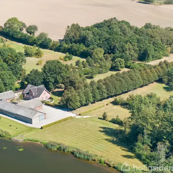 Ejendom, set oppefra, ligger i landlige omgivelser med en stor mark og skov i nærheden. Bygningen er omgivet af træer og græsplæner, og der er en sø i forgrunden.
