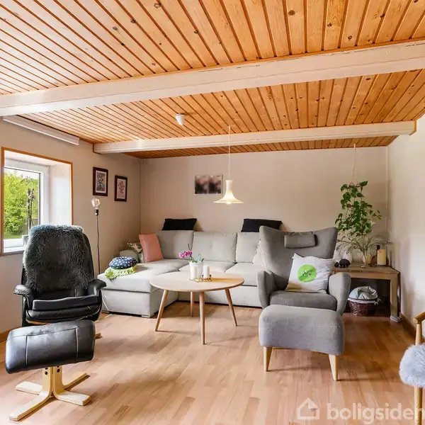 Sofaen står i hjørnet af en lys dagligstue med træloft. Ved siden af står en lænestol. Rummet er dekoreret med planter og små borde, og der er store vinduer.