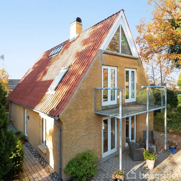 En to-etagers murstenshus med rødt saddeltag har en balkon med glasrækværk. Det står i en have med brostensbelægning, træer og buske i efterårsfarver.