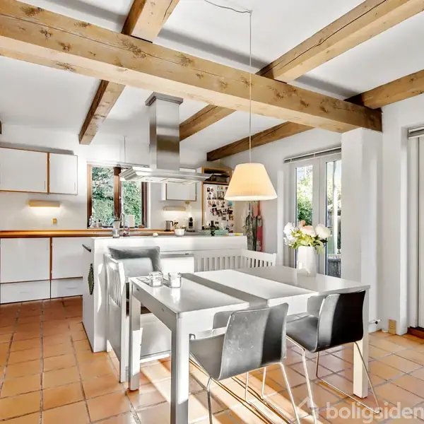 Et moderne køkken med spisebord og sorte stole under en stor lampe. Store vinduer og døre giver udsigt til en terrasse. Loftsbjælker og flisegulv skaber en rustik atmosfære.