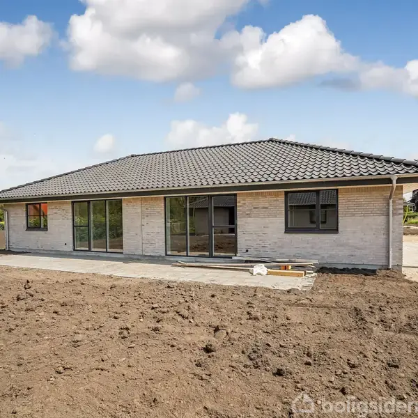 En étplans murstenshus står stille på en nyligt tilsået grund under en blå himmel med spredte skyer. Omkring huset ses andre bygninger og grønt løv i baggrunden.