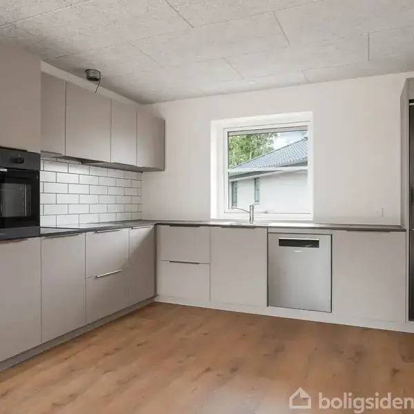 Et moderne køkken med grå skabe og skuffer langs væggene. En indbygget ovn, opvaskemaskine og køleskab er integreret. Et vindue giver udsigt til en bygning udenfor.