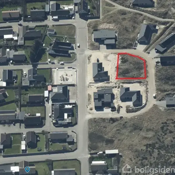 Et boligområde set fra oven, hvor en grund er markeret med en rød linje. Området er omgivet af huse og grønne områder i en kystnær landskab.