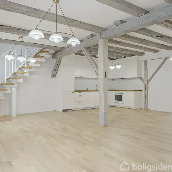 Et køkken med hvide skabe og træbordplade står under en rustik trappe og synlige bjælker. Brune gulvplanker understøtter det lyse og luftige rum. To hængelamper lyser op.