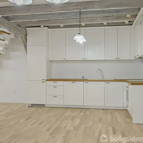 Køkken med hvide skabe og træbordplade står under en trappe i en lys stue. En hængelampe hænger fra loftet, og synlige bjælker præger rummet.