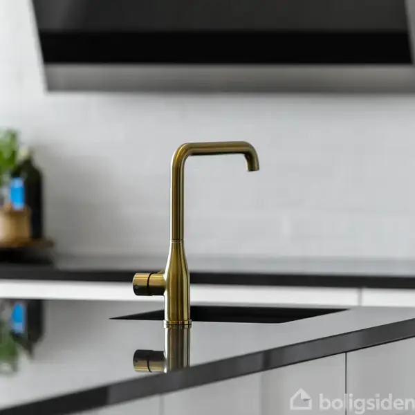 En messingfarvet vandhane står i en moderne køkkenvask. I baggrunden ses krydderiglas og planter på en bordplade. Køkkenet har en minimalistisk, elegant indretning.