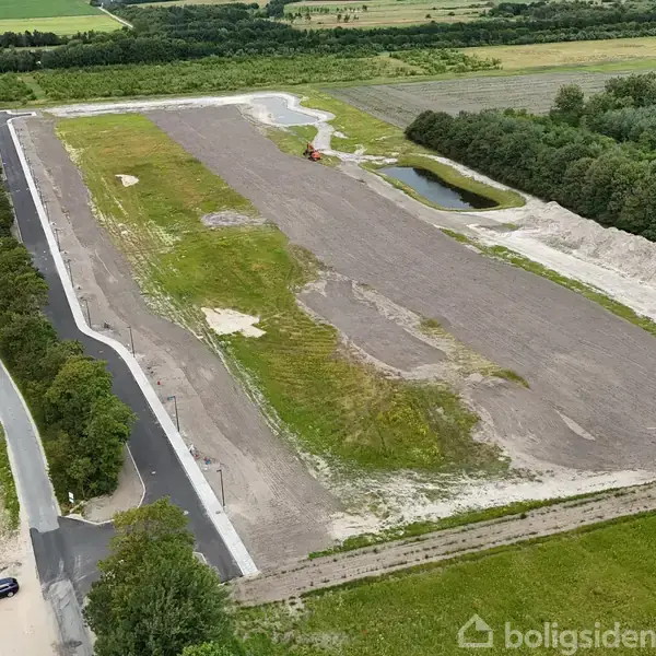 Rektangulært jordareal ligger stille på marker, omkranset af grusveje og en lille bil. Til højre ses en mindre sø ved skovbryn i landligt landskab.