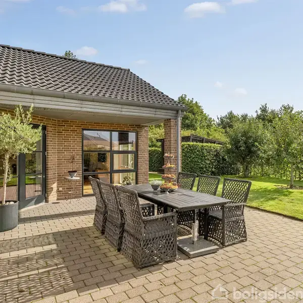 Et udendørs havebord med seks flettede stole står på en flisebelagt terrasse foran en murstensvilla, omkranset af grønne hække og træer og under en klar, blå himmel.