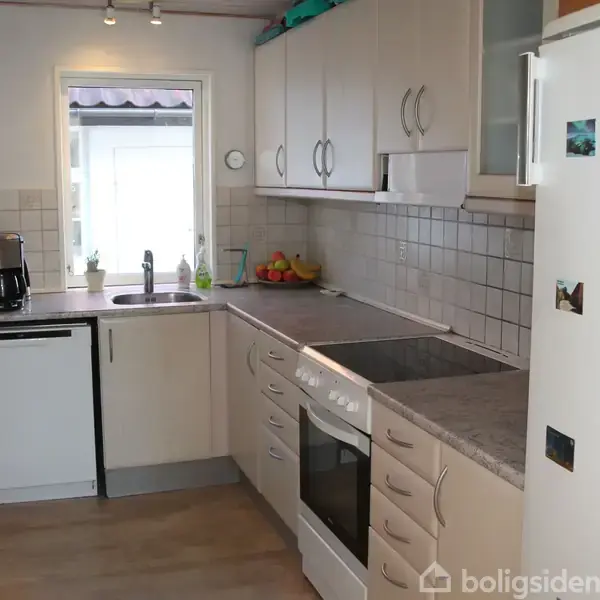 Legetøj fylder reoler og gulvet i et børneværelse med trægulv. Dukkehuse og bamser står ved siden af opbevaringskasser. To sorte stole er placeret ved et lavt bord.