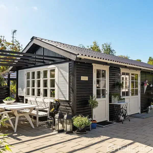 Et sortmalet træhus med hvide vinduer, omgivet af en flisebelagt terrasse med havemøbler. Sollys skinner ned, og omgivende grønne planter giver en hyggelig atmosfære.