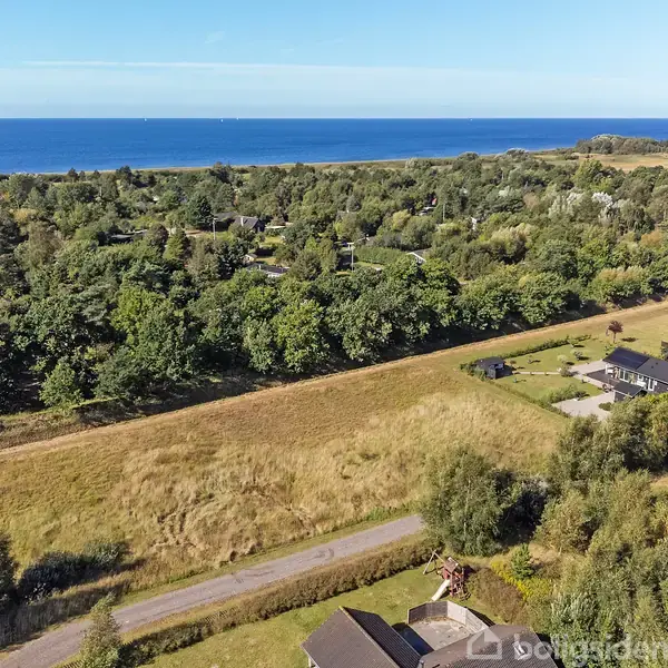Huse ligger spredt i et grønt landskab nær en vej, omgivet af marker og skov med udsigt til en blå havhorisont.