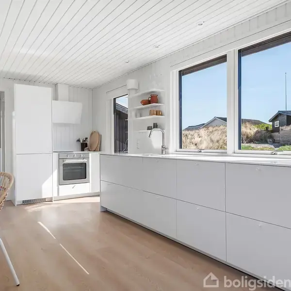 Et hvidt køkken med indbygningsovn og vask. Store vinduer giver udsigt til græs og huse. Rattan stol ved bordet, under et hvidt, træloft.