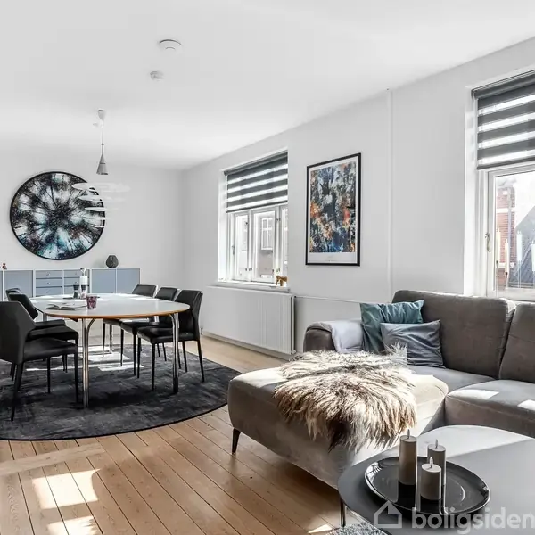 Stue med grå sofa, dækket spisebord og moderne indretning; store vinduer slipper lys ind, og en rund vægdekoration hænger over en skænk. Ingen synlig tekst.