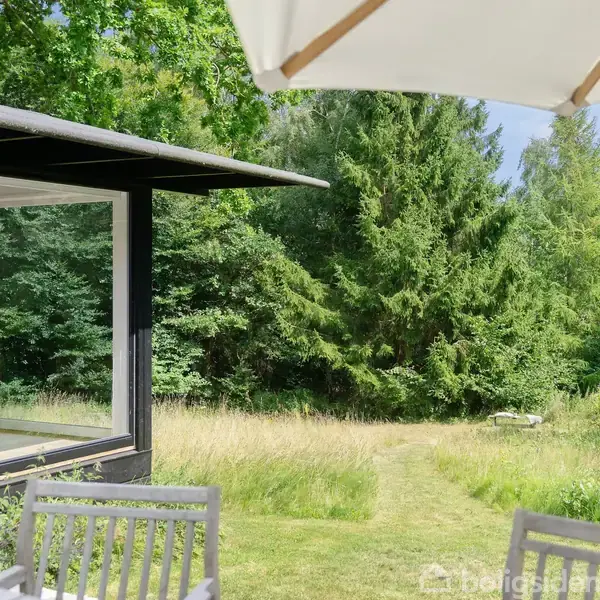 En moderne terrasse med træmøbler og parasol vender ud mod en frodig have med højt græs og tæt skov. Et stort vindue afslører en lysindret stue.