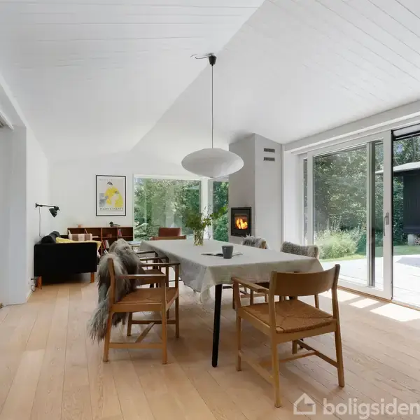Et langt spisebord med stole står i et lyst rum med trægulv og store vinduer. En moderne lampe hænger over bordet, og der er udsigt til en grøn have.