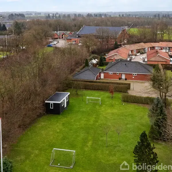 En grøn græsplæne med to fodboldmål står foran et sort havehus. Omkring er der buske og træer, mens man i baggrunden kan se rækkehuse og åbne marker.